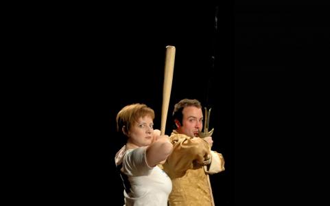 A woman, her body facing sideways but looking at the camera poses with elbows up holds a bat straight up near her face. A man right in front of the woman holds a sword in the same position
