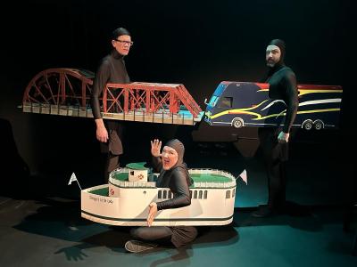 Two people dressed in black in a black room are facing each other expressionless. The one on the left is wearing a bridge that was built around their waist.  The one on the right is wearing a colorful bus built around their waist. Sitting below them on the floor is another person wearing all black with a white boat built around their waste is smiling and waving to the camera.