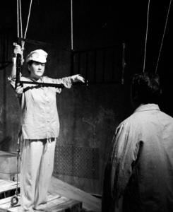 A woman in a plaster military-esque hat peers through a window suspended by ropes. A man stands facing her with his back to the camera.
