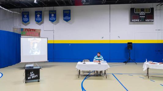 A lone man is sitting at a table inside the gymnasium of middle school. To his right is a screen that has a picture of Richard the Third being projected on it. The gymnasium has banners of accolades on the walls and a scoreboard.
