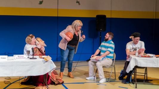 On the left we see a woman who looks as if she is a queen from the 15th Century is seated holding the severed head of a man in hands and she is laughing hysterically. Standing next to her is a blond woman holding a basketball and a microphone and she is yelling at the seated man next to her, a man who looks a bit tight laced. Seated next to him behind a table with strawberries on it is a dejected looking man wearing a black beret.