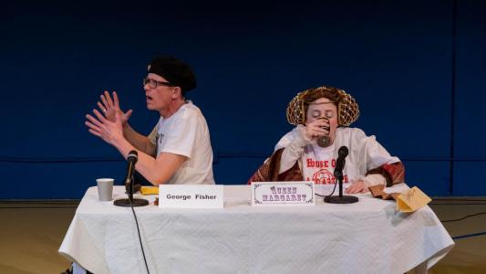 Seated behind a table with two microphones on it are a woman dressed in 15th century clothing with a giant t-shirt over it and a man wearing a black beret and a t-shirt with Richard the Third on it. The man, who has a name tag that reads George Fisher in front of him, is gesturing as if he is trying to explain something while the woman, who has a name tag that reads Queen Margaret in front of her, is drinking wine out of a cup.