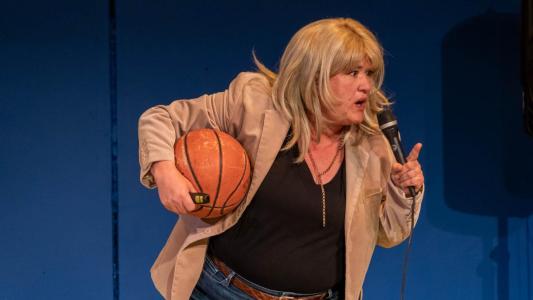 A blond woman in a blazer is holding a basketball under her left arm. She has a microphone in her right hand with her index finger pointed. She looks as if she is giving someone a stern talking to.