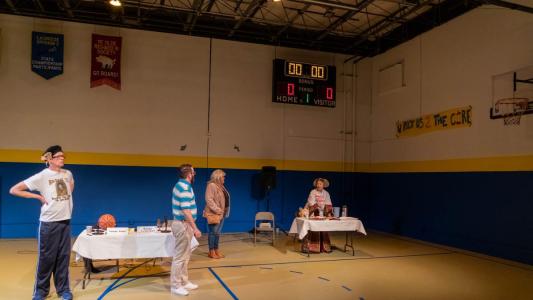 Four people are standing on what looks like a middle school basketball court in a gymnasium. On the wall above a woman that looks like she is from the 15th Century is a score board that reads zero/zero. On the wall next to her is a basketball hoop and hand painted sign that reads “You rock us to the core”. Standing next to her on the basketball court is another woman and 2 men and a table with 2 microphones on it.  Above them, on the wall behind them there are two banners that read “Lacrosse Division Three State Championship Participants” and “Ye Olde Richard the Third Society. Go Boars.”