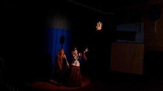 Two women, one dressed as Queen Margaret, stand in front of a blue curtain. It is dark around them except for the severed head she has just tossed into the air like a basketball.