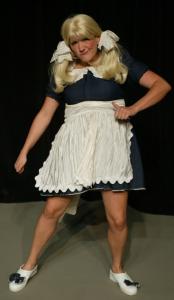 A woman in blond pigtails makes a silly face while she dances. She wears a dark blue dress with a white collar and apron.