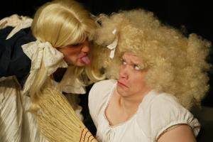 Close up of a woman in a long, blond wig sticking her tongue out at a very scared Cinderella who is holding a broom up for protection.