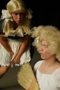 A woman in blond pigtails leans over blowing a raspberry in the face of Cinderella, who looks upset and holds a broom.