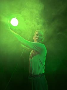 In a smoky space, a man reaches up to adjust a theatrical light. The light casts a green glow across the picture and picks up the details of the smoke.