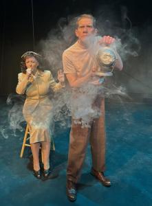 A man holding an old fashioned fog machine stands in the foreground spraying fog towards the picture taker. A woman in 1940's garb sits behind him on a stool coughing.