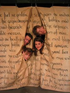 Four people poking their heads through the backdrop with the Old English Poem Beowulf written on it.