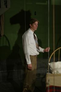 A man in a white shirt and striped tie stands hunched holding a wire that is suspending a bed above the floor. Behind him is the shadow of a person in a camel costume.