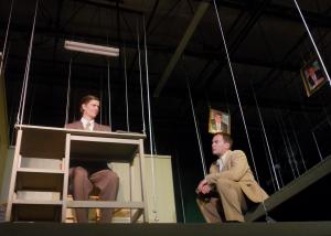A man in a brown suit is sitting at a small desk suspended above the floor by wires. He is smiling at another man who is seated facing him on a wire suspended walkway and wearing a tan suit with no tie. There are two identical photos of a man with black hair’s face hanging above.