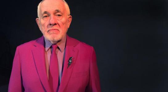 an older bearded man in a bright fuchsia suit looks sternly at the camera. He is surrounded by darkness and has a lightning bolt lapel pin.